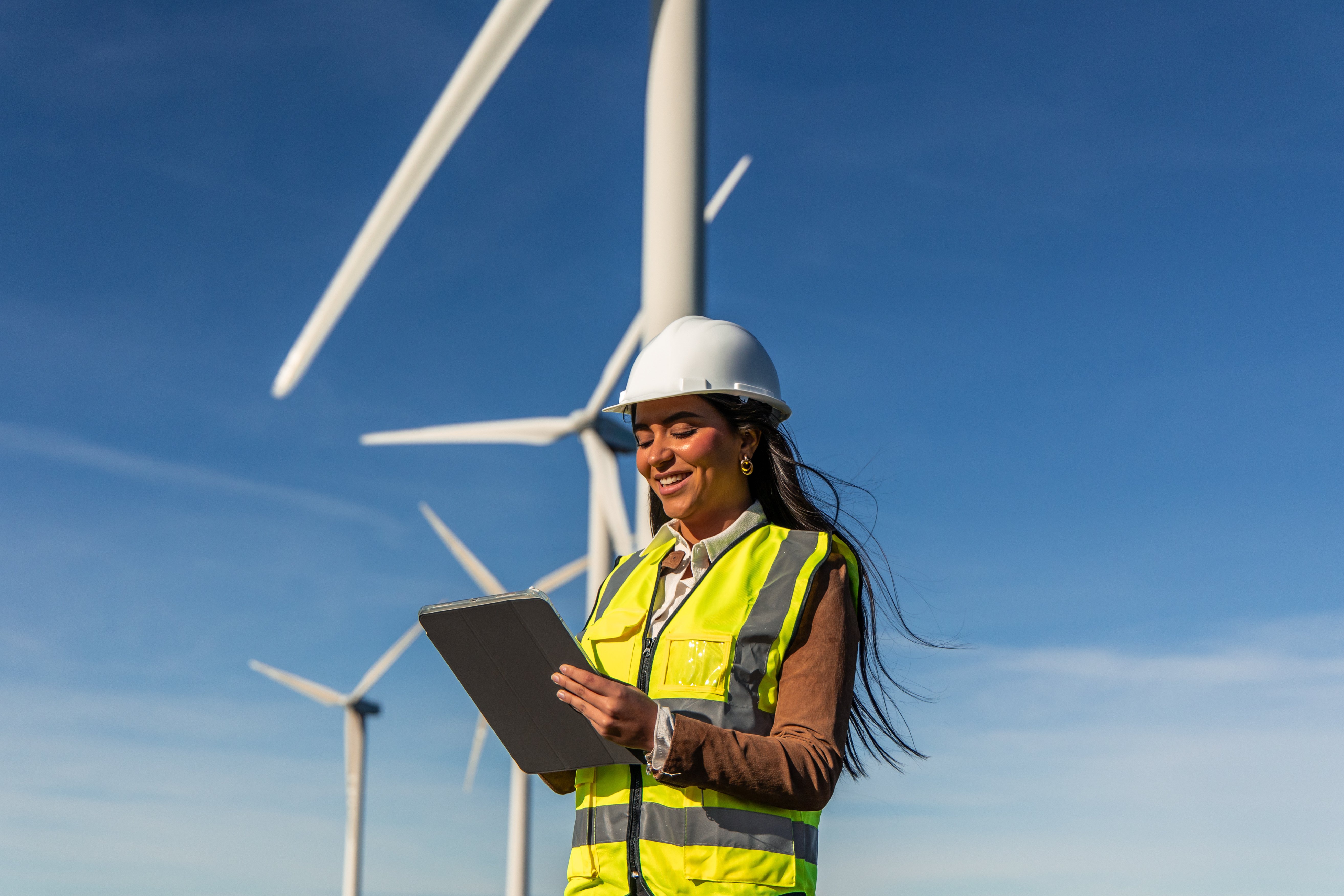 Female engineer in hi-vis and hardhat reading a clipboard in front of windturbines with a blue sky